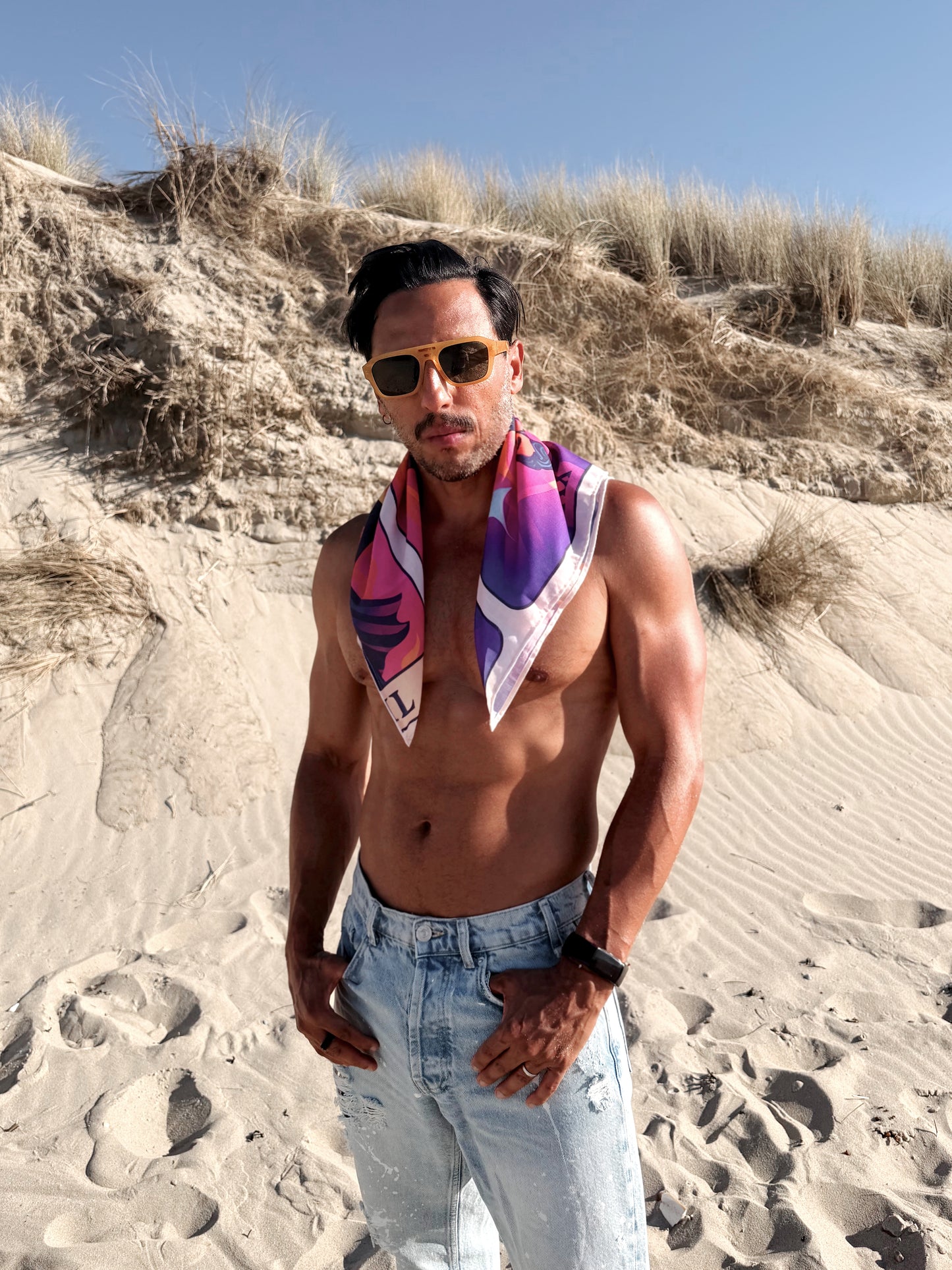 Man standing on a sandy beach wearing sunglasses and a colorful scarf