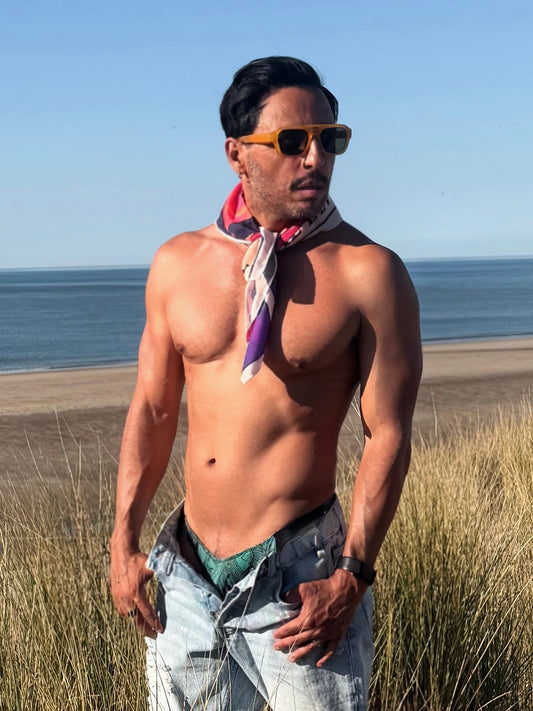 Man standing on a beach wearing sunglasses and a colorful bandana The Devil, with ocean and sky in the background.