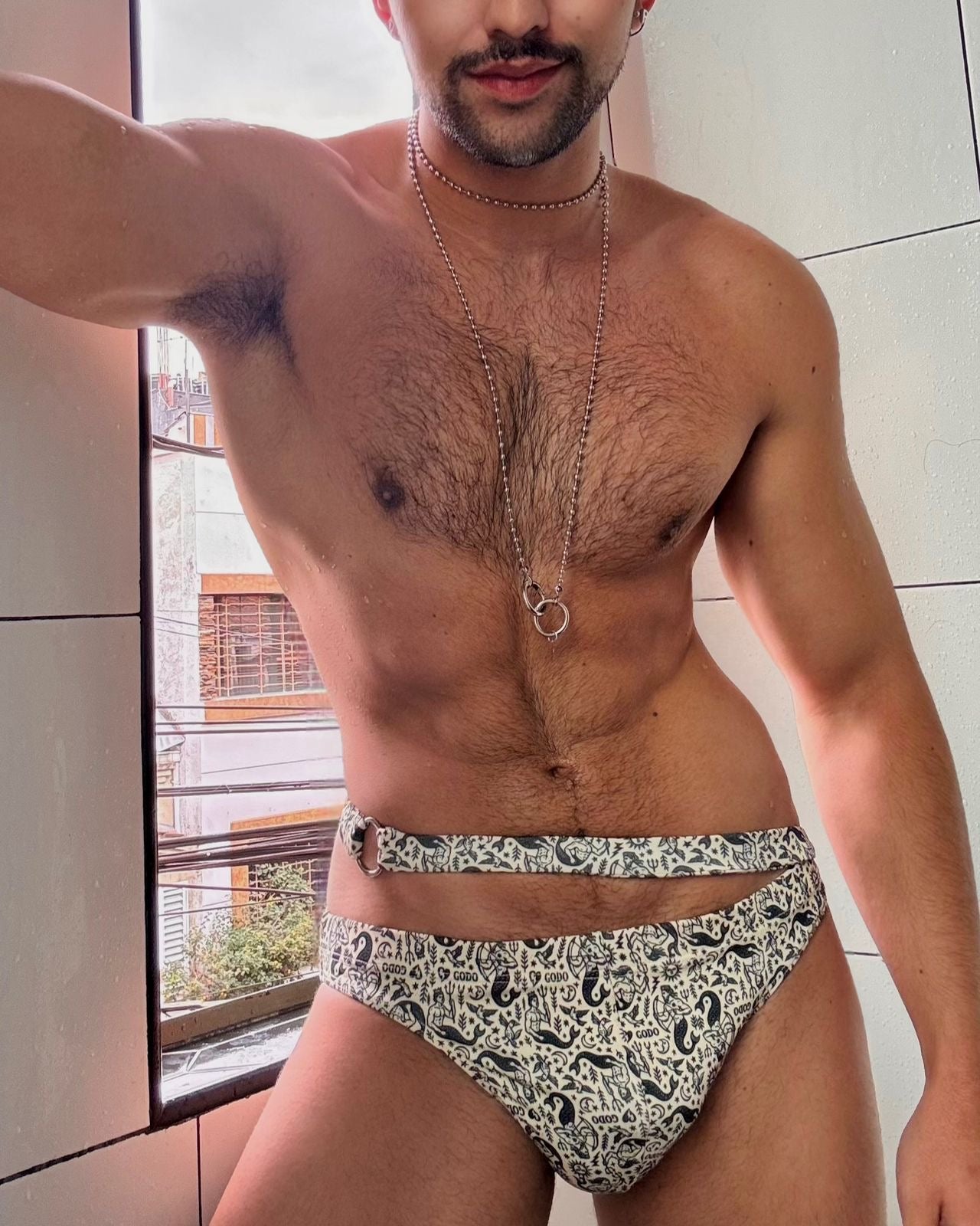 Man wearing a patterned swimsuit standing in front of a window.