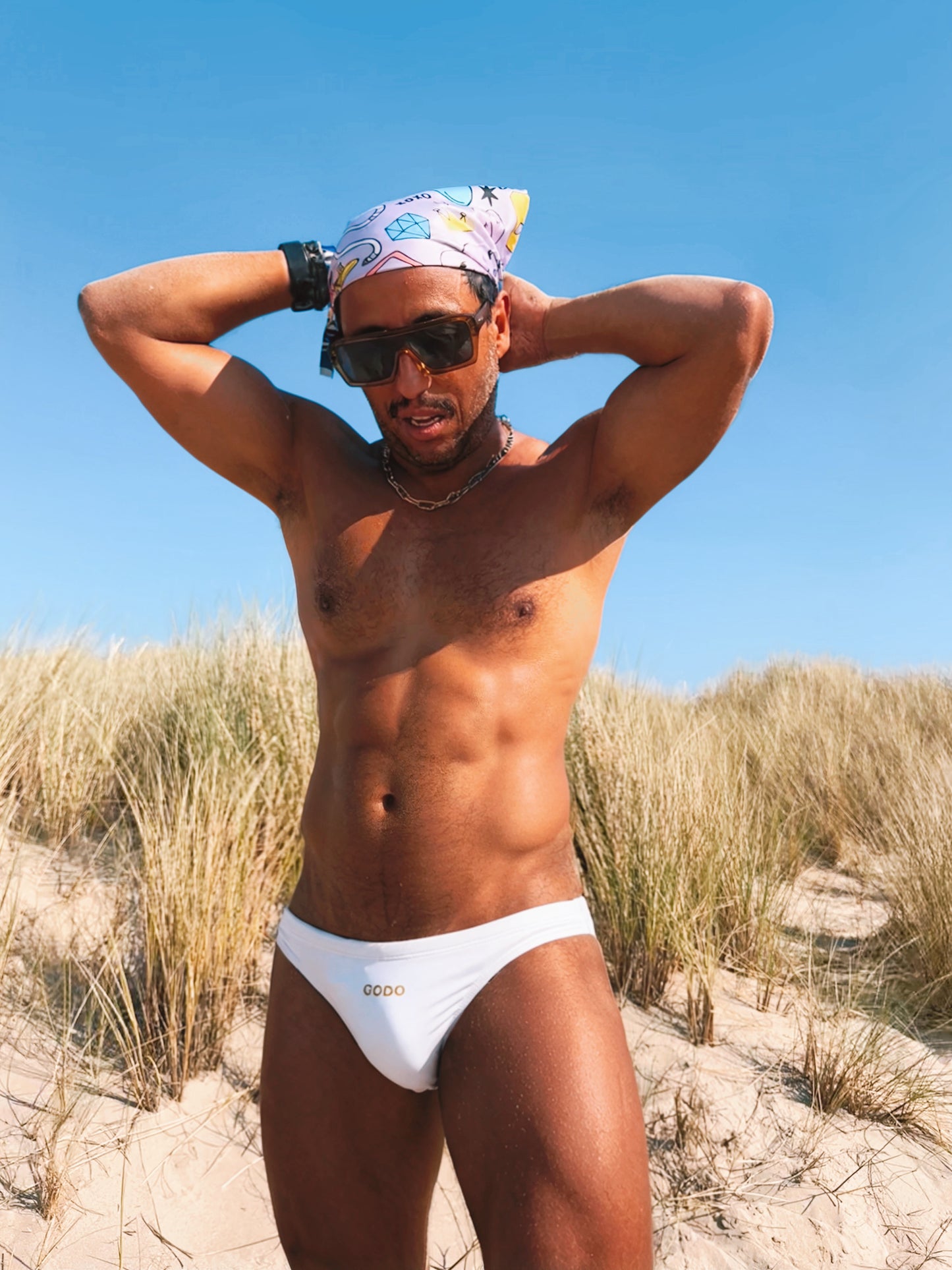 Man in white bikini bottom and colorful headscarf standing on a sandy beach with clear blue sky.