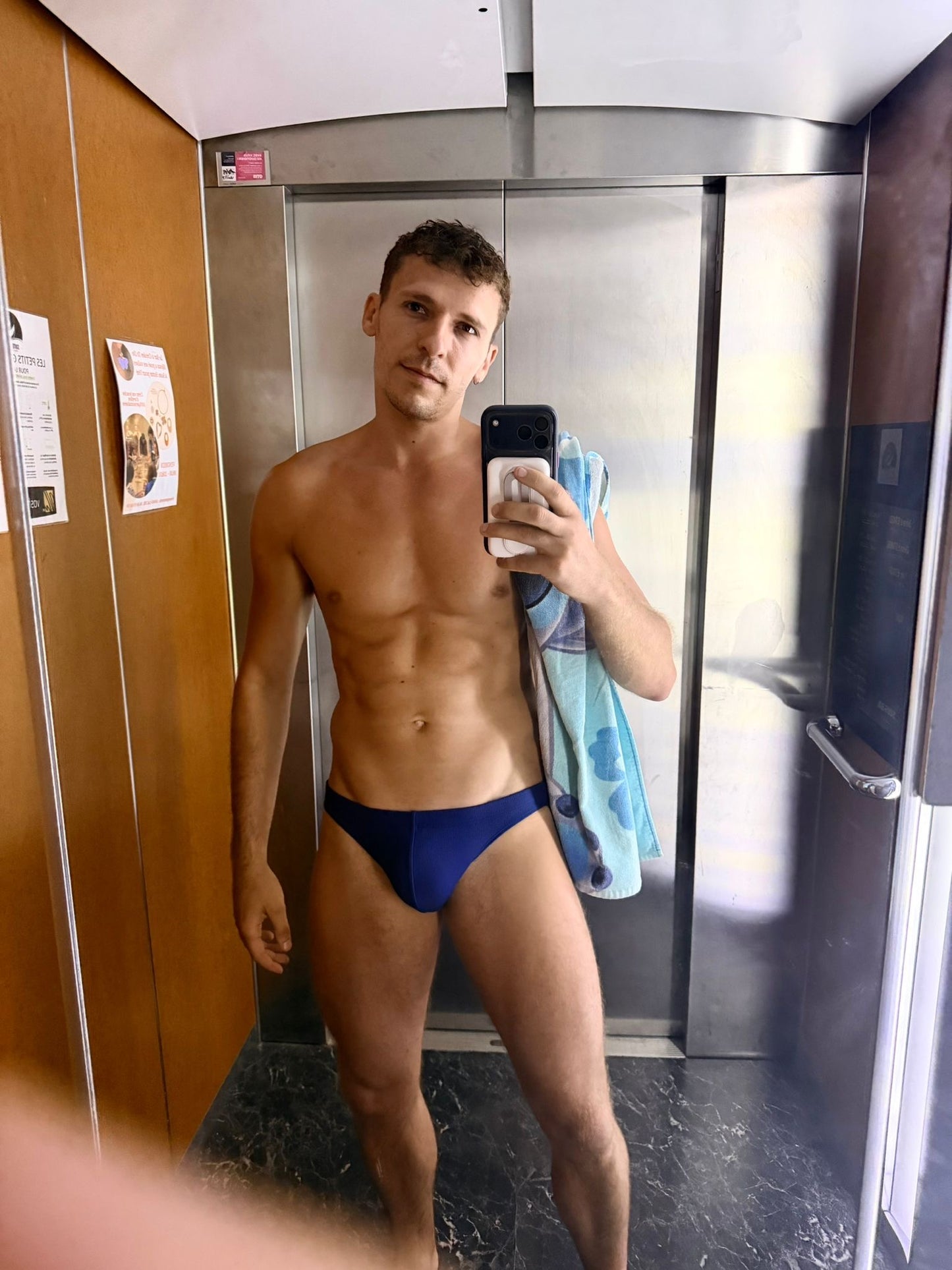 A man taking a mirror selfie in an elevator, wearing a dark blue swim brief and holding a light blue towel over his shoulder.