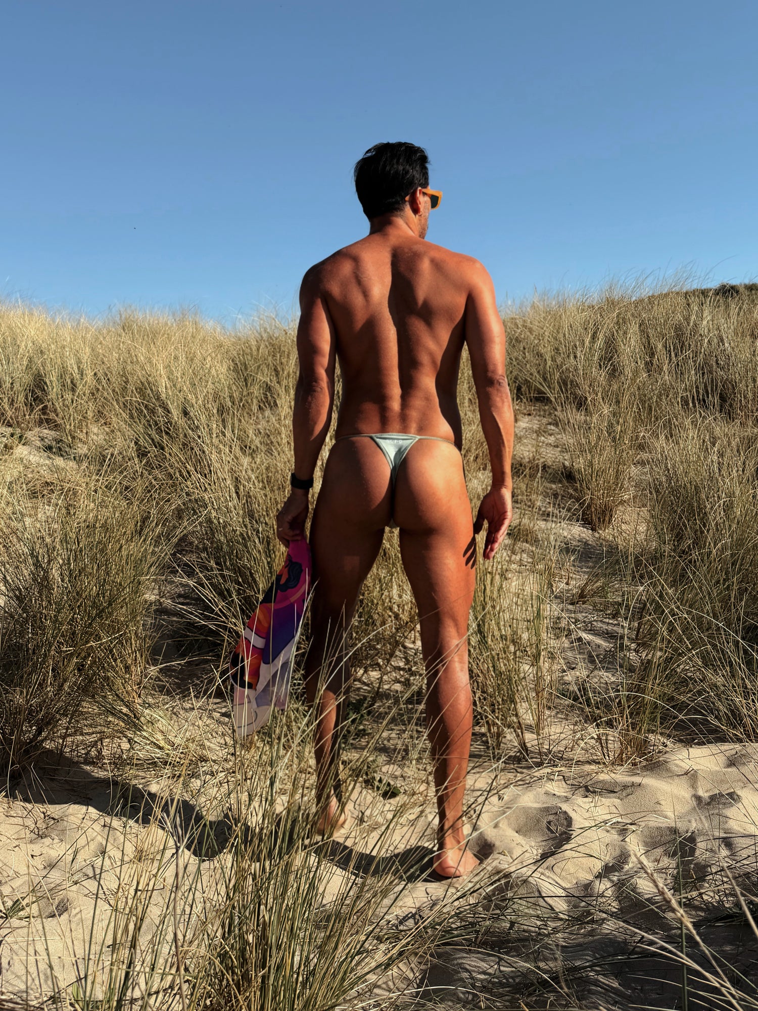 Man walking on a sandy beach with dunes, holding a colorful bandana.