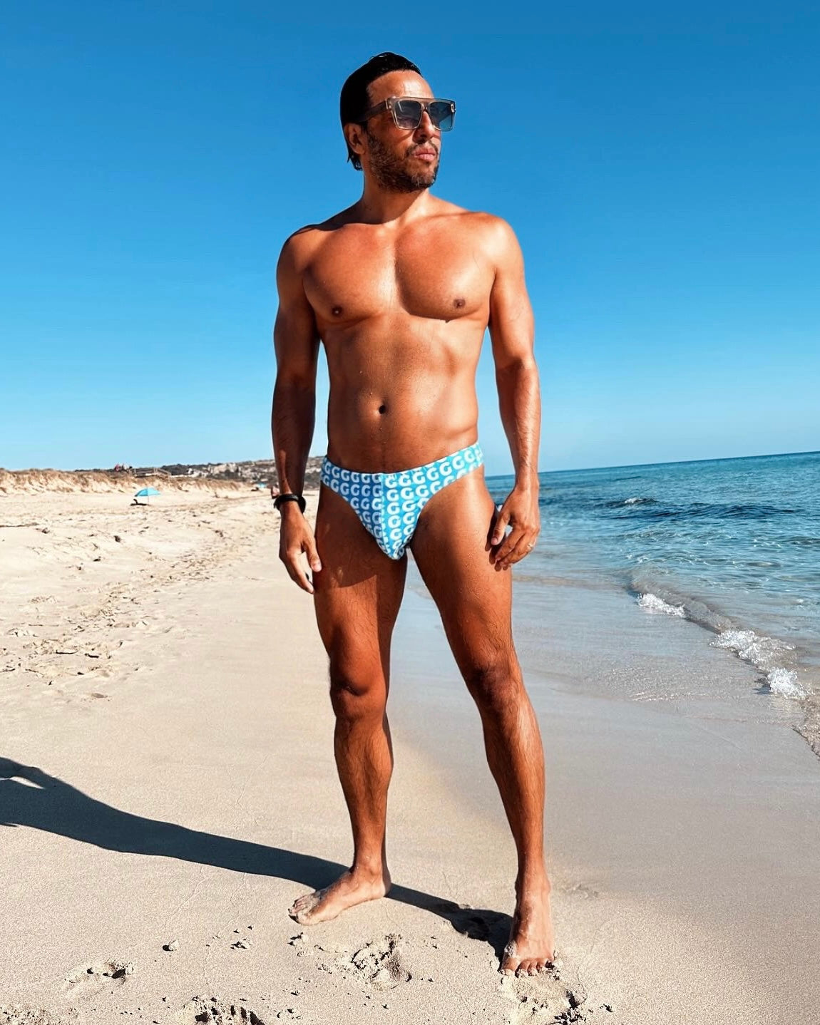 Man in a blue patterned swimsuit standing on a beach with clear blue sky and ocean.