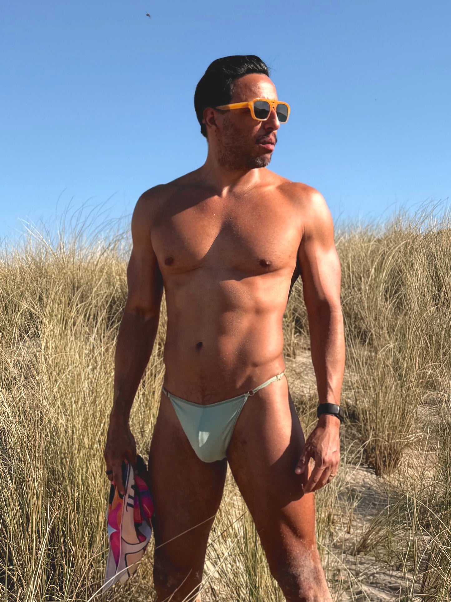 Man in light-colored swim trunks standing on a beach with sand dunes and clear sky.