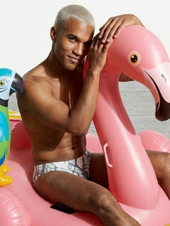 Man wearing a snake swimbrief sitting on a pink inflatable flamingo pool float with a colorful parrot float in the background.
