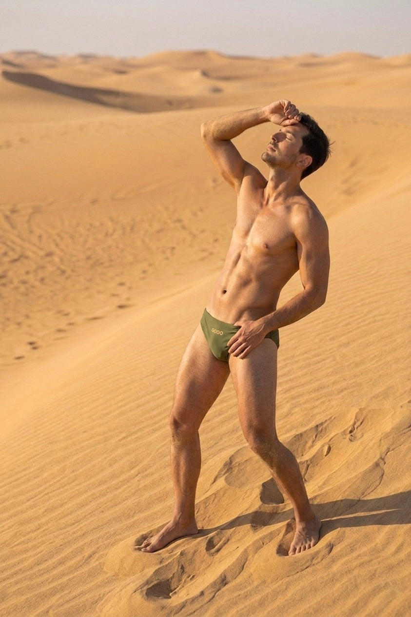 Man standing in a desert wearing green swim trunks.