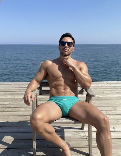 Man in teal swim shorts sitting on a wooden chair by the ocean