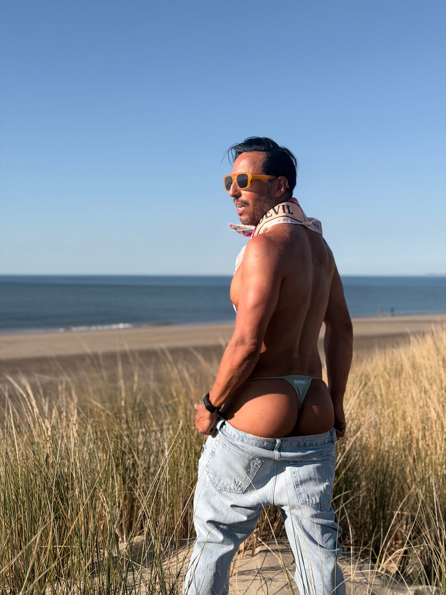 Man showing his thong while standing on a beach with ocean view wearing sunglasses and a bandana around his neck.