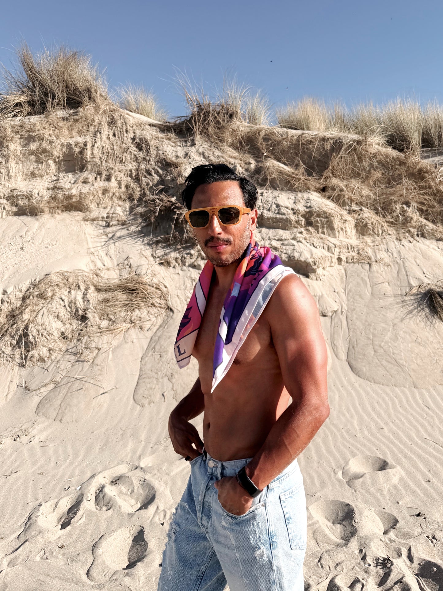 Man standing on a sandy beach wearing sunglasses and a colorful bandana.