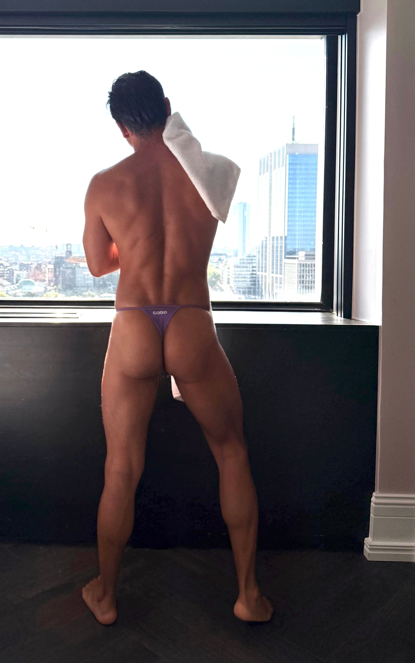Man standing by a window with a cityscape view, wearing a purple thong.